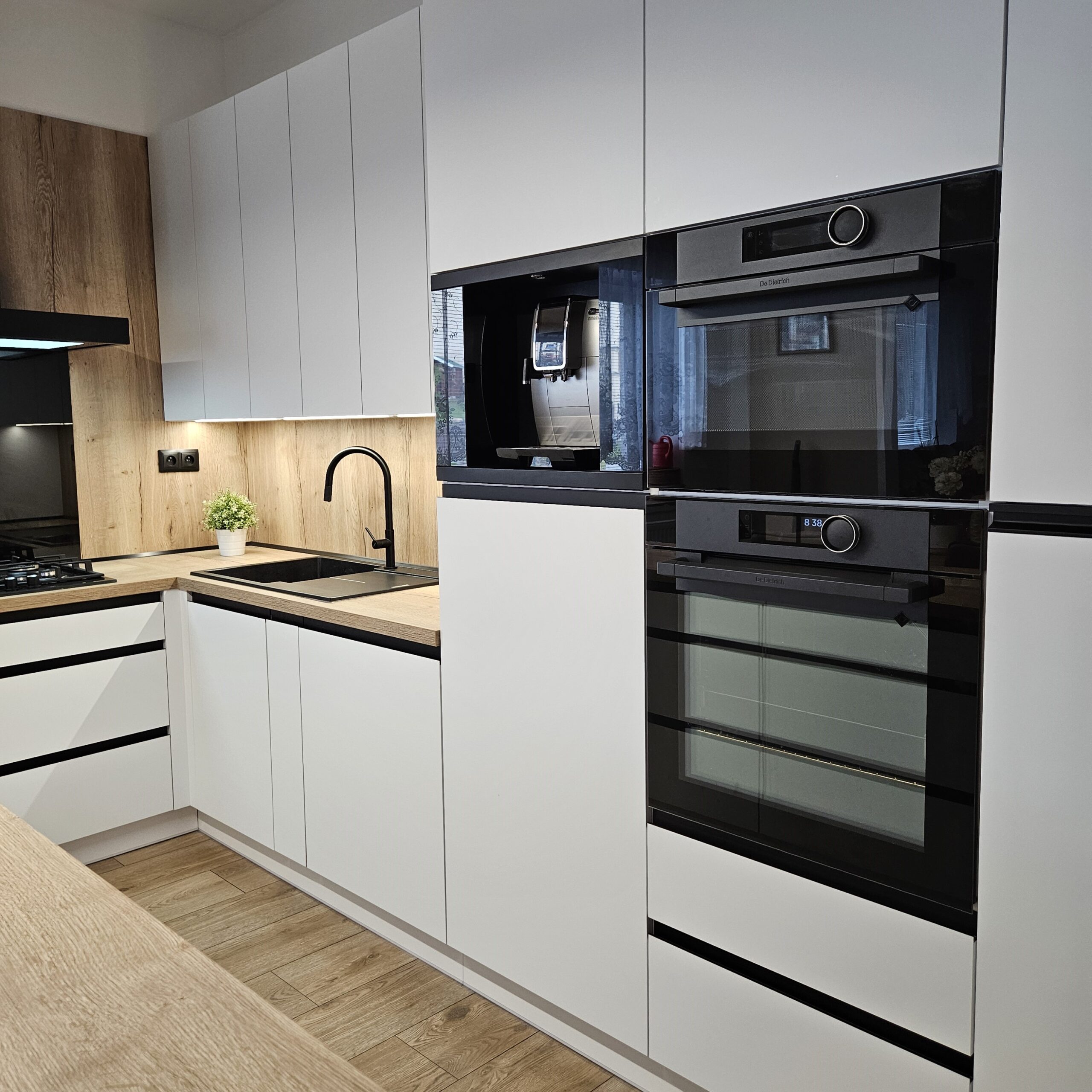 Elegantní kuchyň na míru pro klientku na Smržovce Místo, kde design neustupuje pohodlí. Tato kuchyň spojuje precizní řemeslo s moderními prvky, čímž vzniká harmonický prostor, který je stejně krásný jako funkční. Korpus v odstínu Arktická bílá doplňuje pracovní deska a zadní obklad v dekoru Dub Halifax přírodní, které dodávají interiéru útulnost a přírodní charakter. Spodní skříňky jsou osazeny zápustnými úchytkami Grande v elegantní černé matné, horní části pak využívají subtilní profil S068 v identickém odstínu pro zachování čistých linií. Samozřejmostí je kvalitní kování rakouské firmy Blum, které zajišťuje dlouhou životnost a pohodlné každodenní používání. Kuchyň je dále vybavena luxusními spotřebiči De Dietrich.