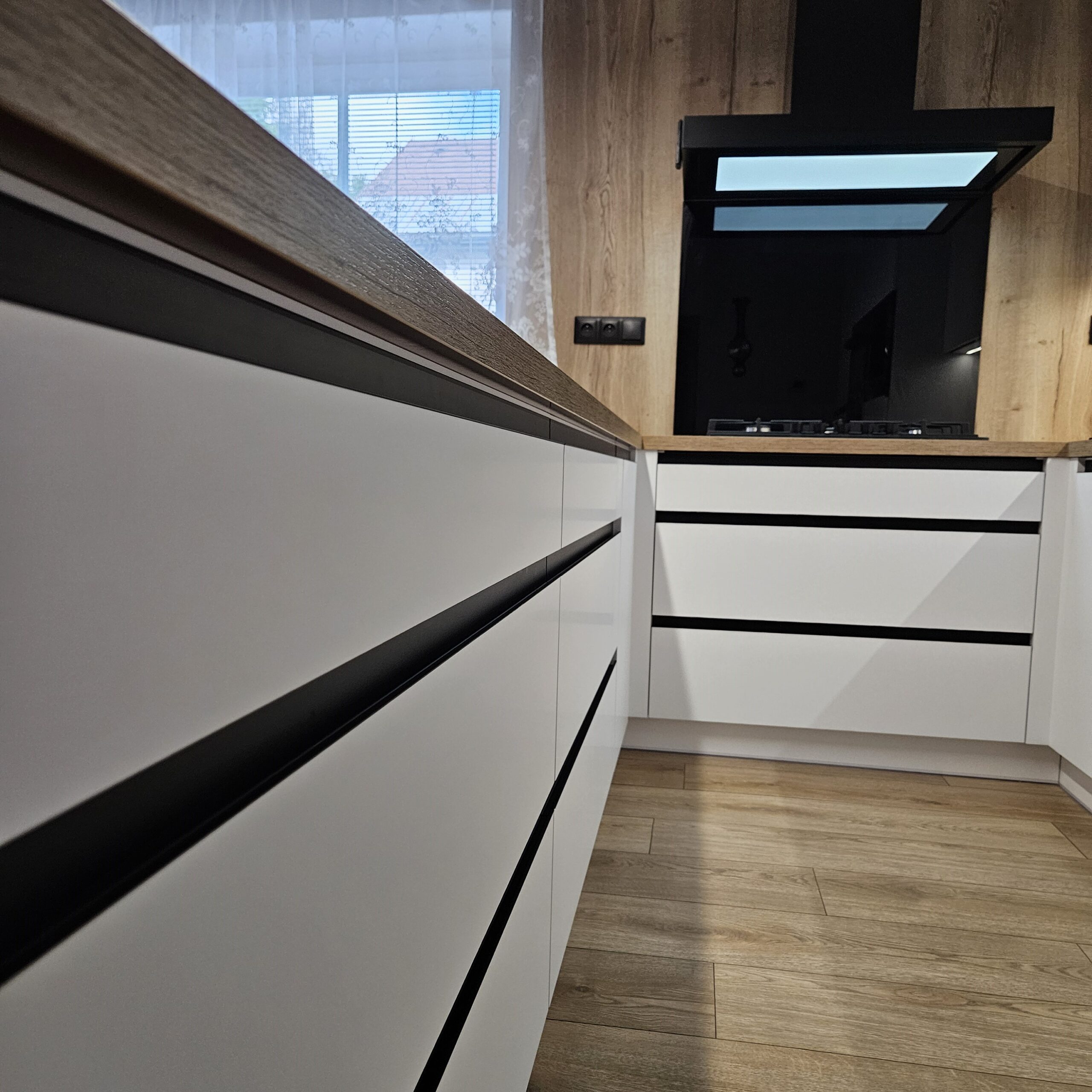 Elegantní kuchyň na míru pro klientku na Smržovce Místo, kde design neustupuje pohodlí. Tato kuchyň spojuje precizní řemeslo s moderními prvky, čímž vzniká harmonický prostor, který je stejně krásný jako funkční. Korpus v odstínu Arktická bílá doplňuje pracovní deska a zadní obklad v dekoru Dub Halifax přírodní, které dodávají interiéru útulnost a přírodní charakter. Spodní skříňky jsou osazeny zápustnými úchytkami Grande v elegantní černé matné, horní části pak využívají subtilní profil S068 v identickém odstínu pro zachování čistých linií. Samozřejmostí je kvalitní kování rakouské firmy Blum, které zajišťuje dlouhou životnost a pohodlné každodenní používání. Kuchyň je dále vybavena luxusními spotřebiči De Dietrich.